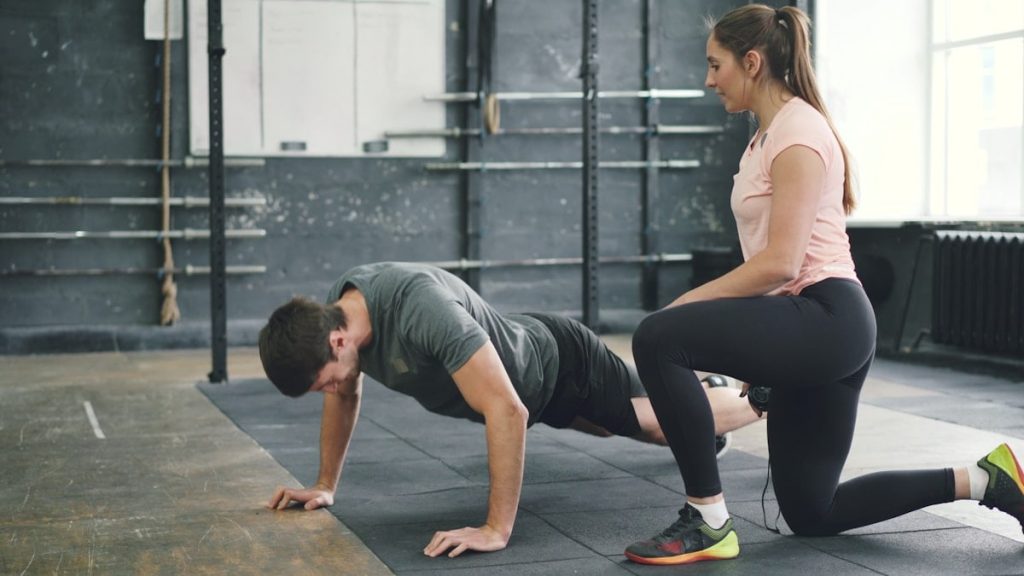 Strong guy is doing push-ups in sports gym while trainer young woman is speaking motivating student. Healthy activity, lifestyle and sports concept.