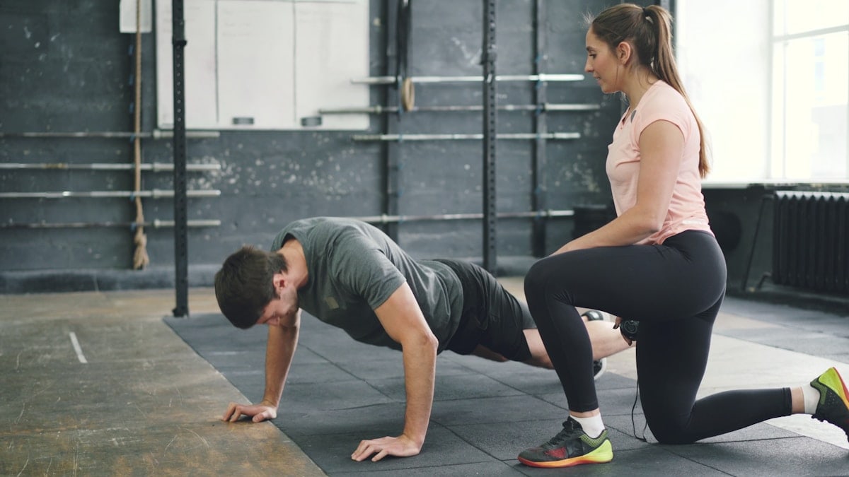 Strong guy is doing push-ups in sports gym while trainer young woman is speaking motivating student. Healthy activity, lifestyle and sports concept.